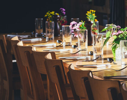Ein Bildausschnitt eines gedeckten, hellbraun farbenen Tisches mit mehreren Stühlen. Der Tisch ist zudem mit bunten Blumen geschmückt und wird mit Licht angestrahlt.